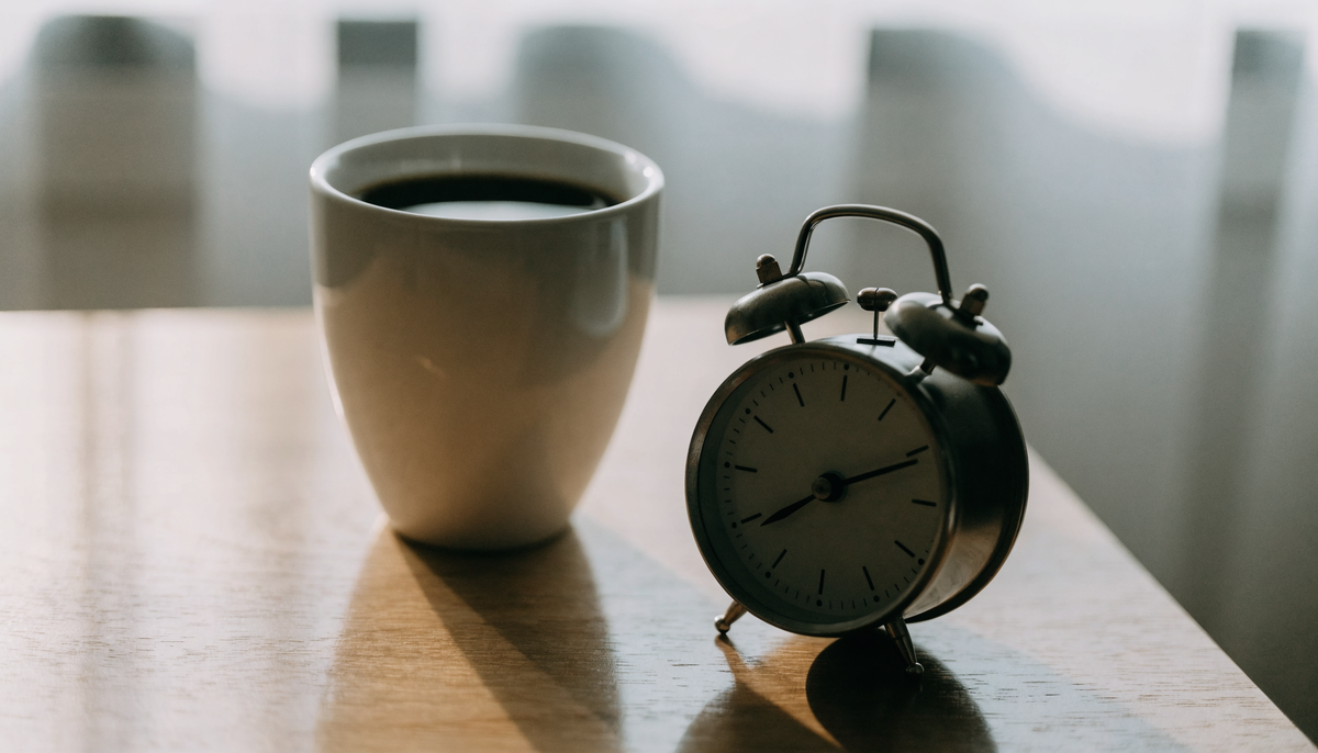 Tasse de café noir posée à côté d'un réveil sur une table en bois clair